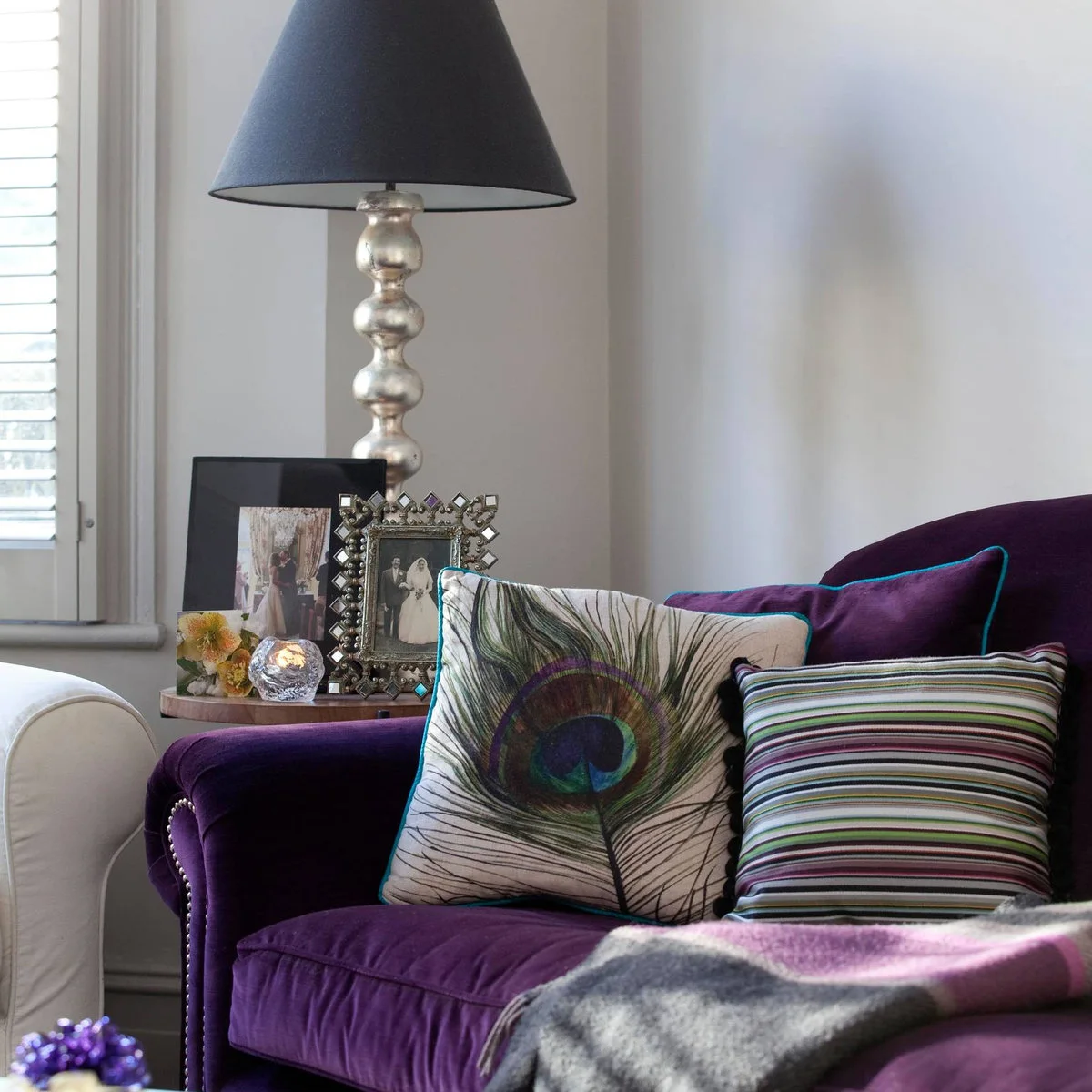 Purple sofa detail with peacock-patterned and striped cushions beside a window and table lamp