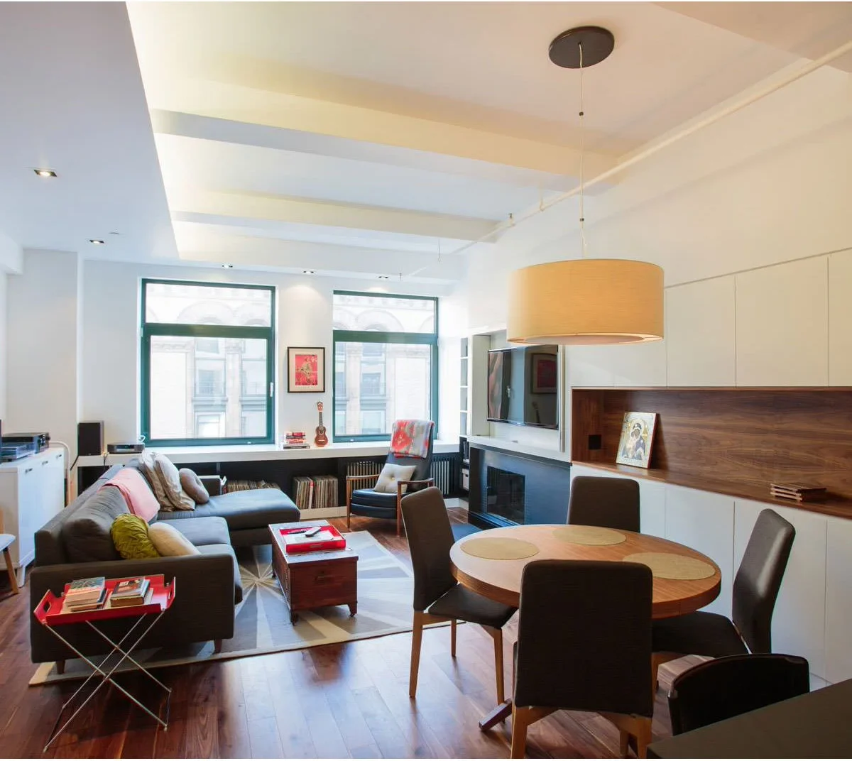 A dark sofa faces a wall television in a narrow room, with a round wood dining table and dark chairs behind the seating area.