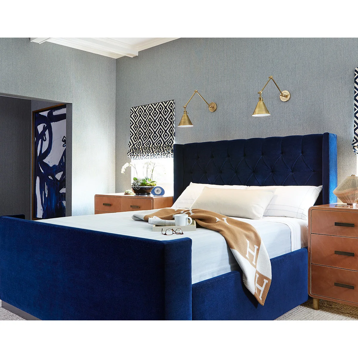 Deep blue upholstered bed against light gray walls with caramel-colored bedside chests and brass wall lamps.