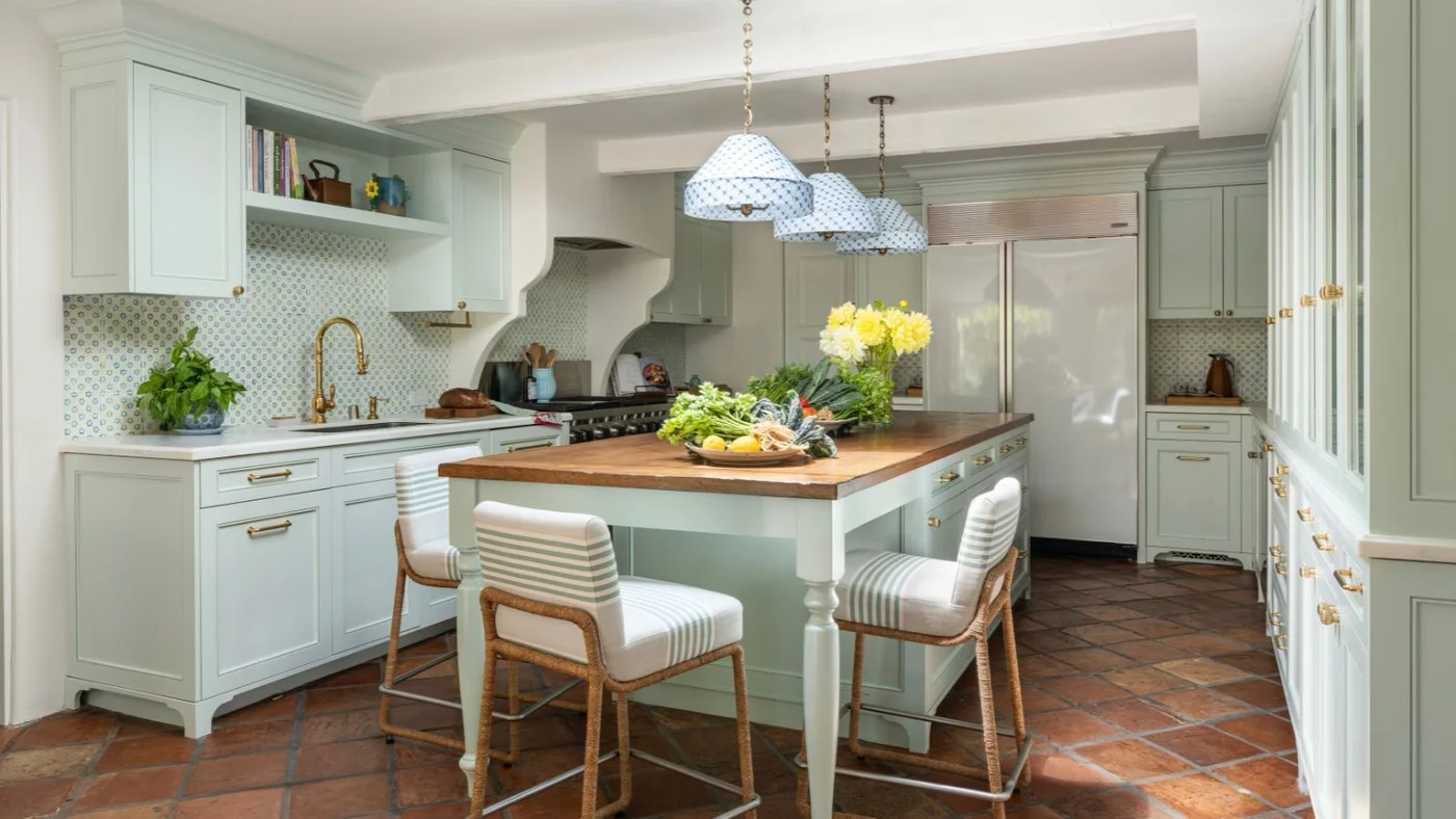 Weathered terracotta squares surround a pale blue-green kitchen island with a timber top and striped stools.