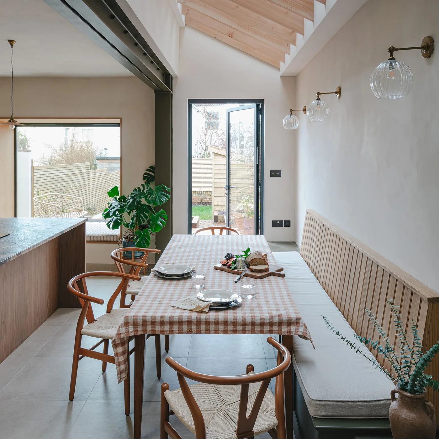 A long dining table with wooden chairs and a cushioned wall bench stands beside a kitchen island and glazed garden door