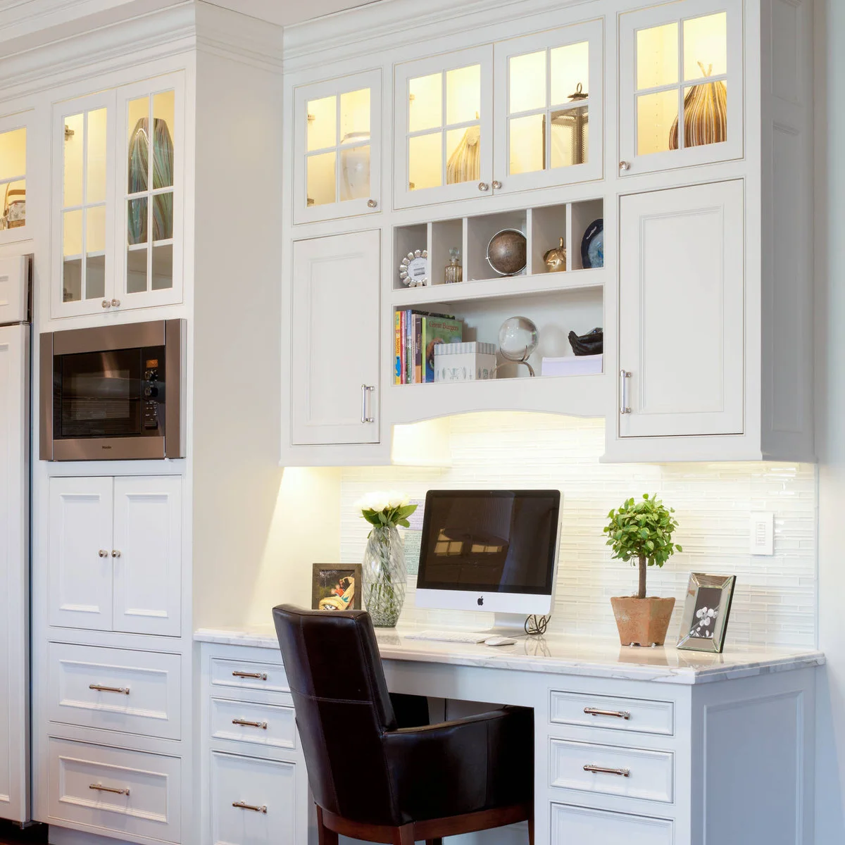 White kitchen-side desk with a monitor, dark chair, drawers and illuminated glass-front cabinets