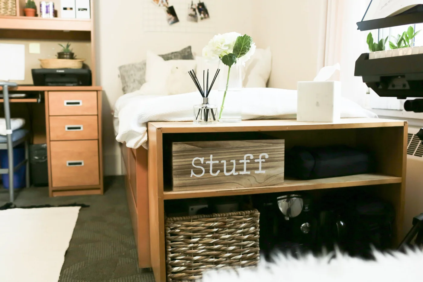 Low wood shelf at the foot of a student bed with a labeled box, woven basket and stored items