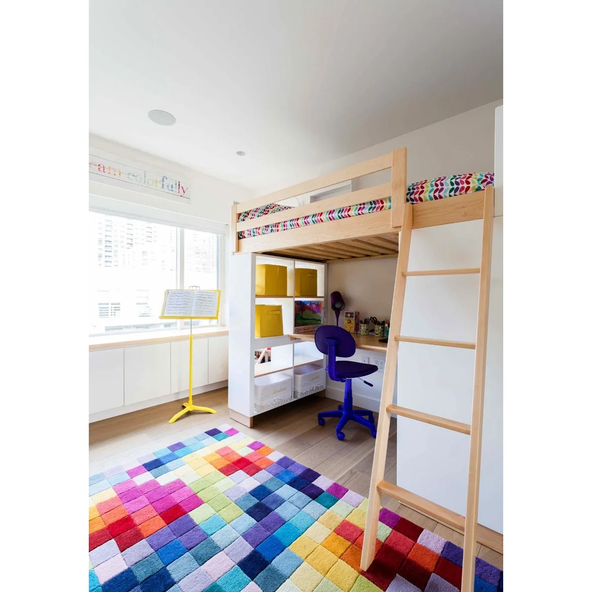 Pale wood loft bed above a desk with a purple chair, white shelves, yellow bins and a multicolored rug.