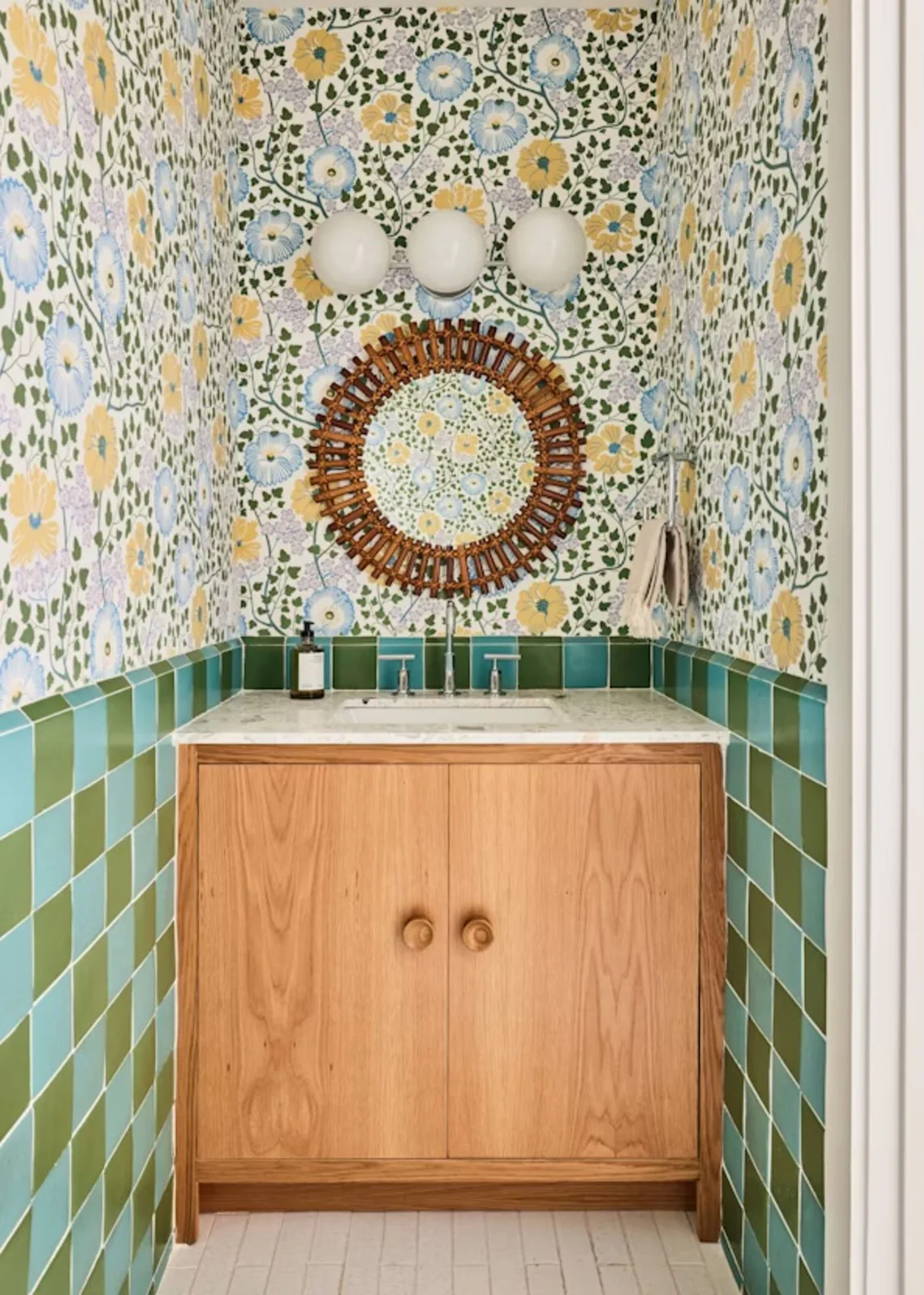 A small bathroom combines yellow-and-blue floral wallpaper, green and blue checked lower tiles, a wood vanity, round woven-look mirror and three white globe lights.