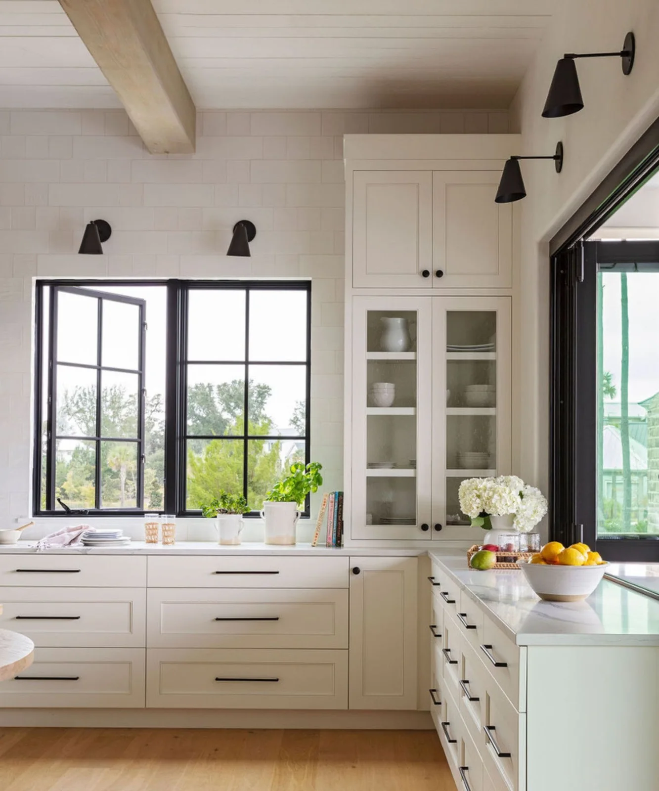 White framed cupboards and drawers with dark handles sit beside dark-framed windows and simple black wall lights.