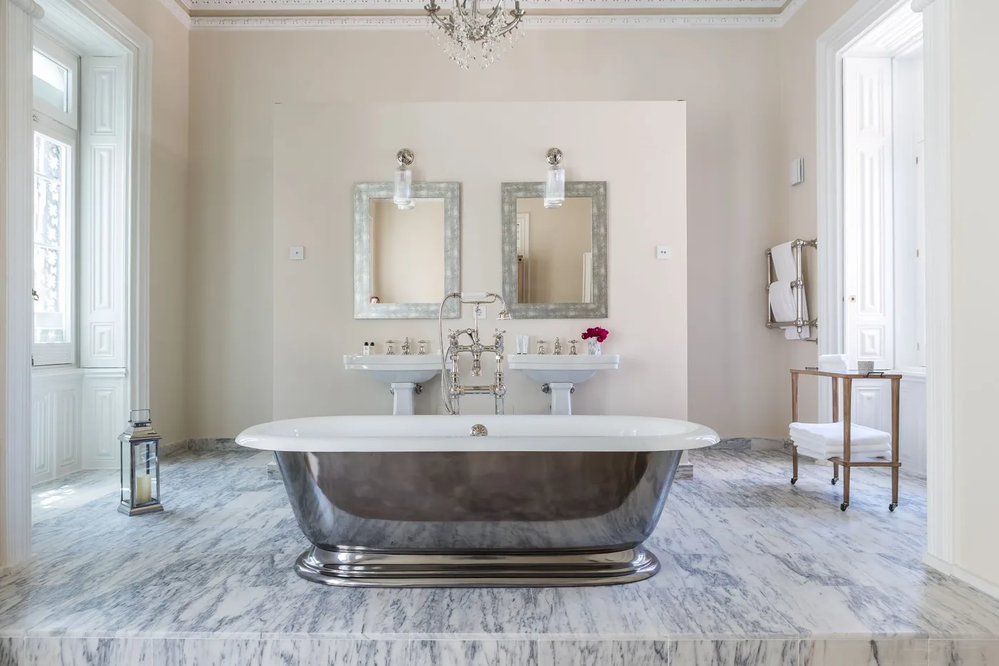 Metal-toned freestanding tub centered before two pedestal basins and framed mirrors in a pale bathroom.