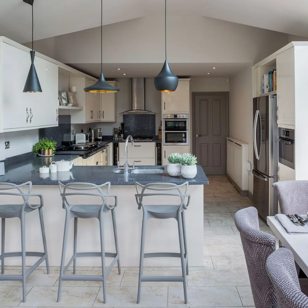 Pale cabinets surround a dark peninsula counter beneath three dark pendants, with creamier fronts deeper in the kitchen.