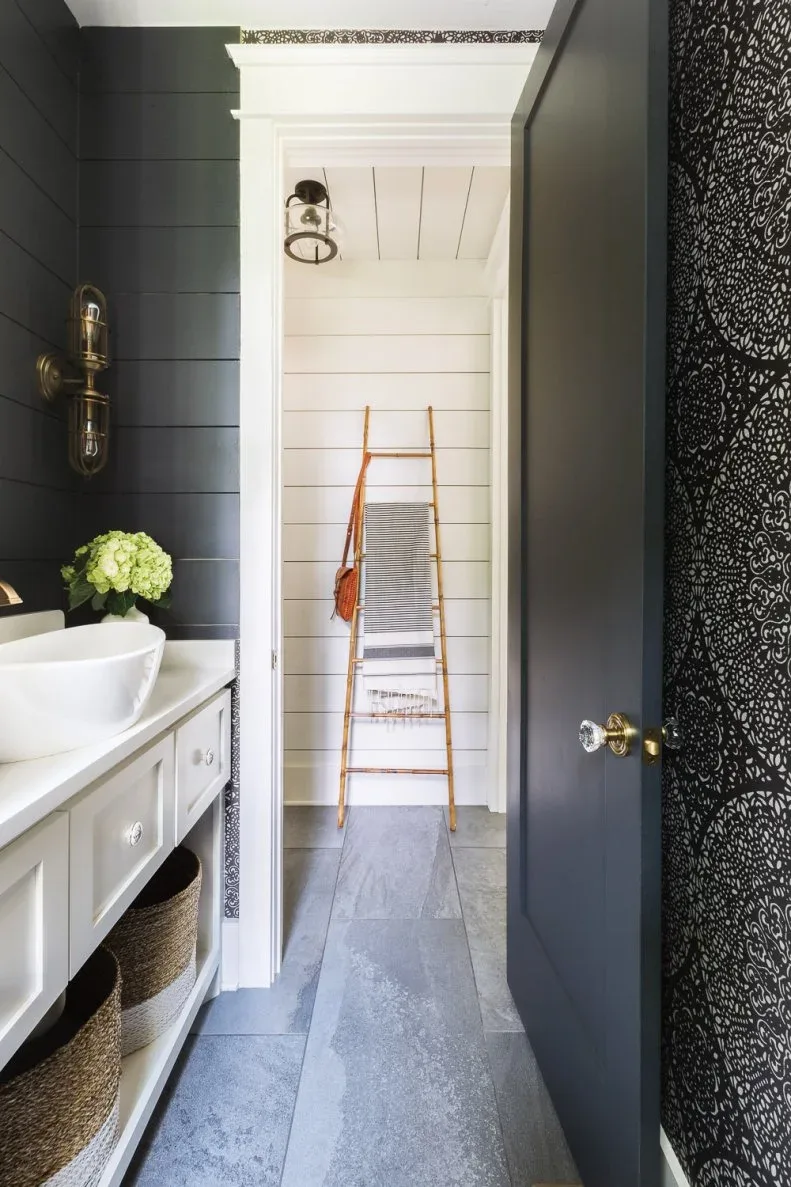 Dark horizontal wall boards surround a white basin and cupboard beside a patterned wall section, with a towel ladder visible through the adjoining doorway