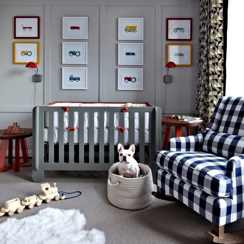 Gray crib against paneled wall with vehicle pictures, checked adult chair, red wall lights and side table.
