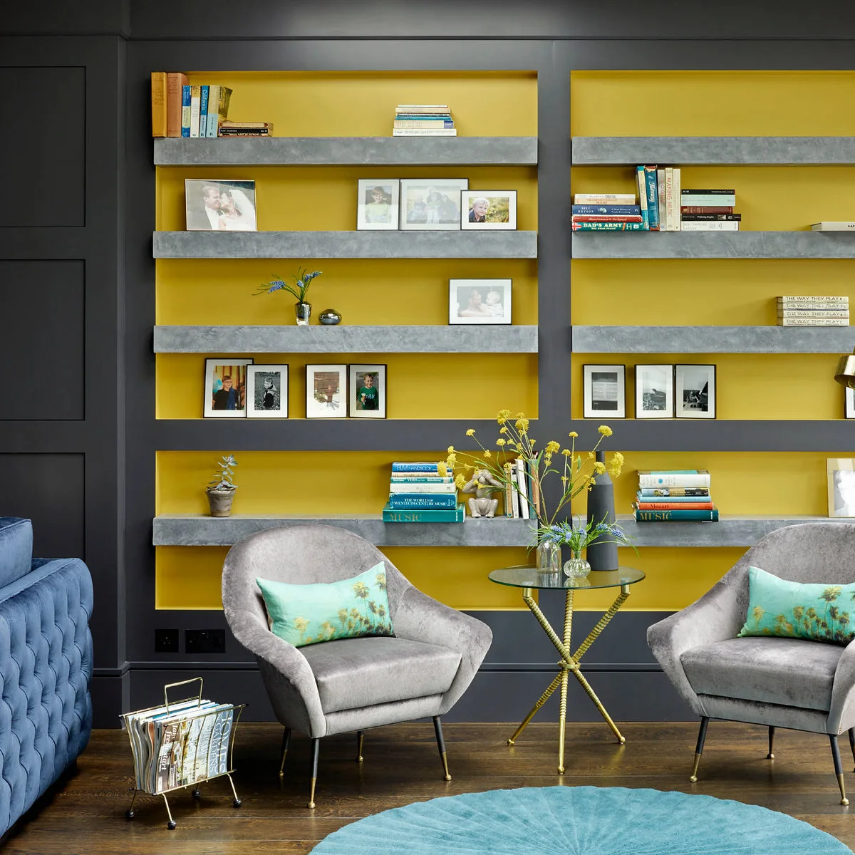 Charcoal-framed shelving with yellow backs above two gray armchairs, a small glass table and blue rug