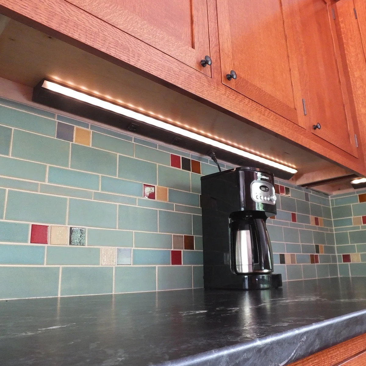 A slim bright light and dark outlet rail run beneath warm wood cabinets above a coffee maker and blue tiled backsplash.