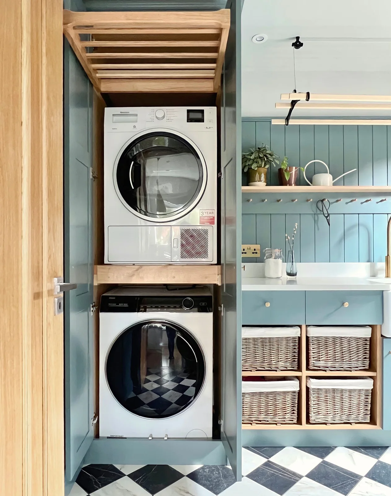A tall wood cabinet holds stacked laundry appliances beside blue paneled cabinetry, a sink, basket cubbies, a peg rail and a suspended drying rack.