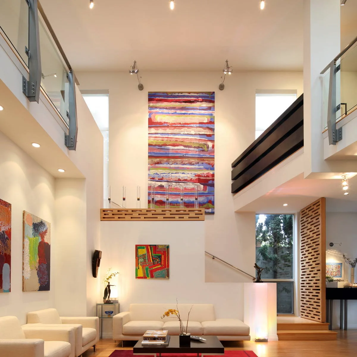 A double-height white living room with colorful vertical artwork, pale seating and upper glass-and-steel walkways.