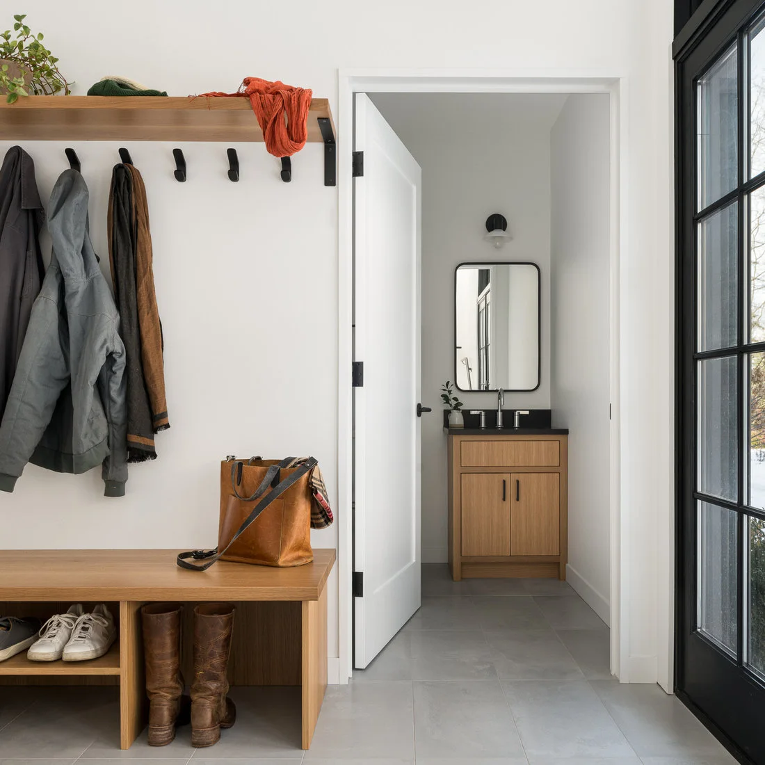 A wood bench with shoe compartments stands below coat hooks and a wood shelf beside a glazed exterior door and a small bathroom doorway.