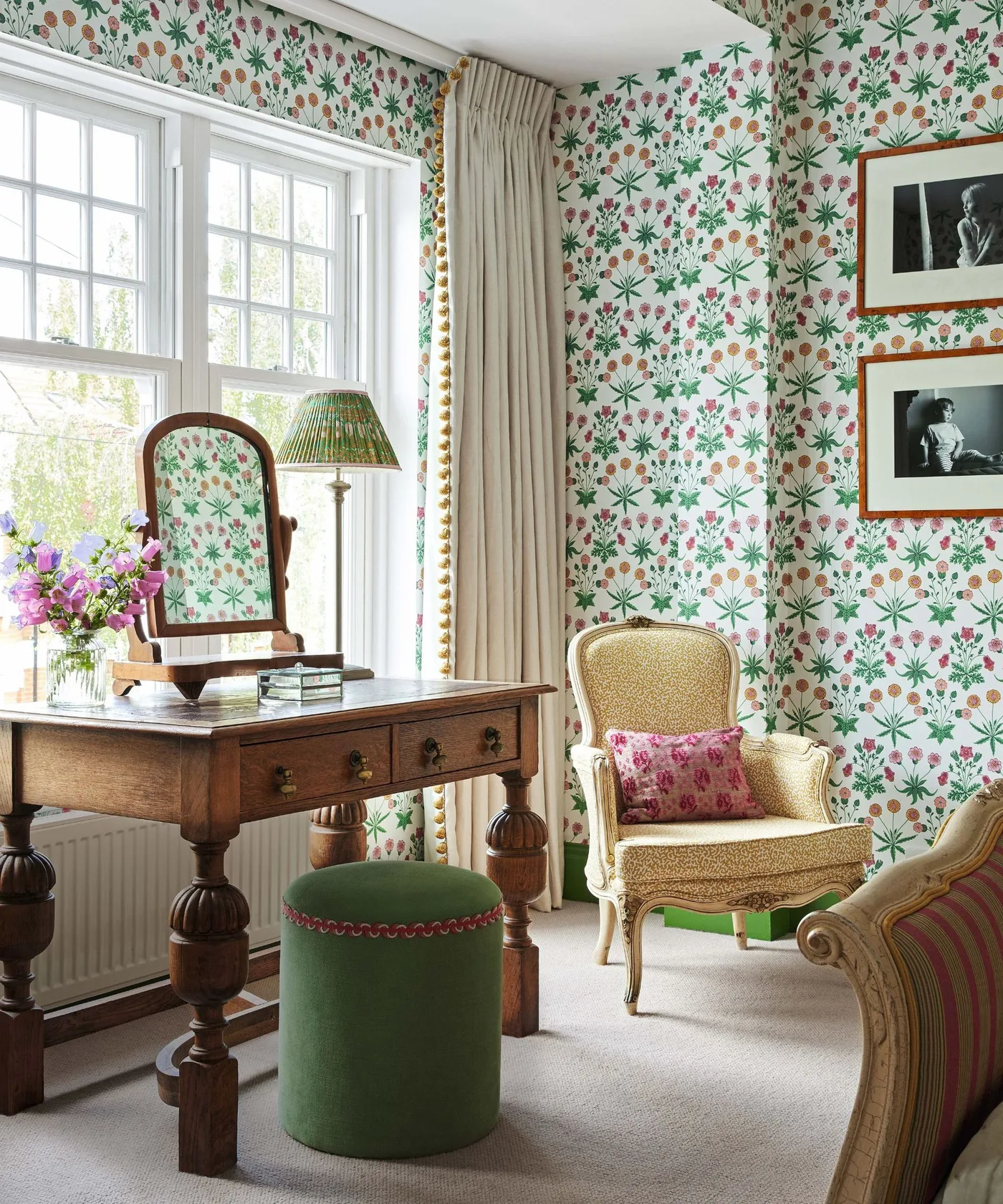 Bedroom dressing corner with floral wallpaper, wooden dressing table and mirror, green stool and upholstered chair beside a window.