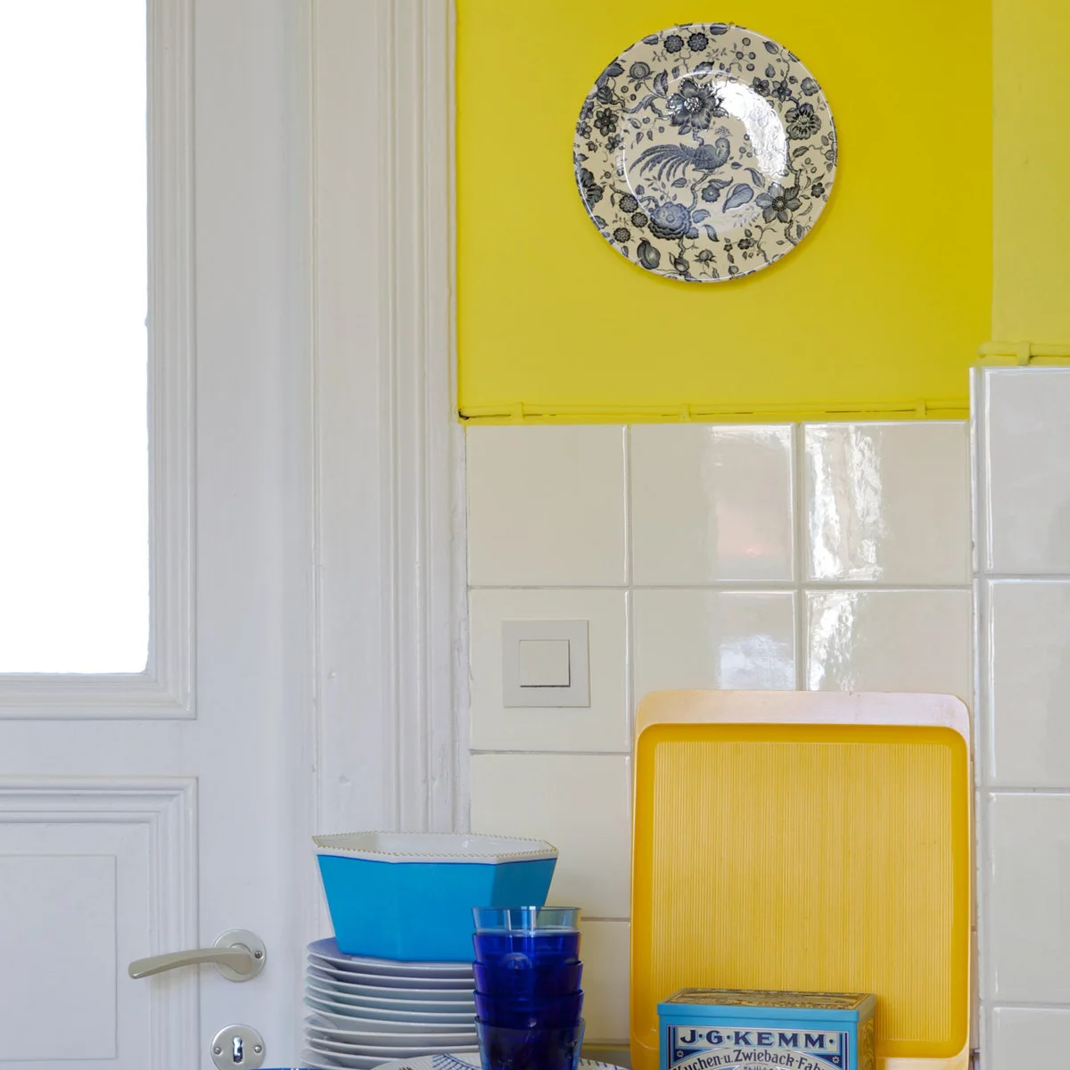 Yellow kitchen wall above white tiles with a yellow tray, blue glasses and stacked dishes