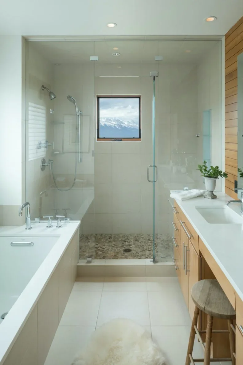 A narrow pale bathroom has a tub on one side, wood-fronted vanity and small stool on the other, and a glazed shower with a window at the end