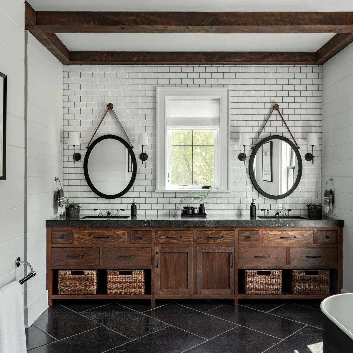 Dark wood double vanity with a black counter, central tray, airy greenery and baskets on the lower shelf.