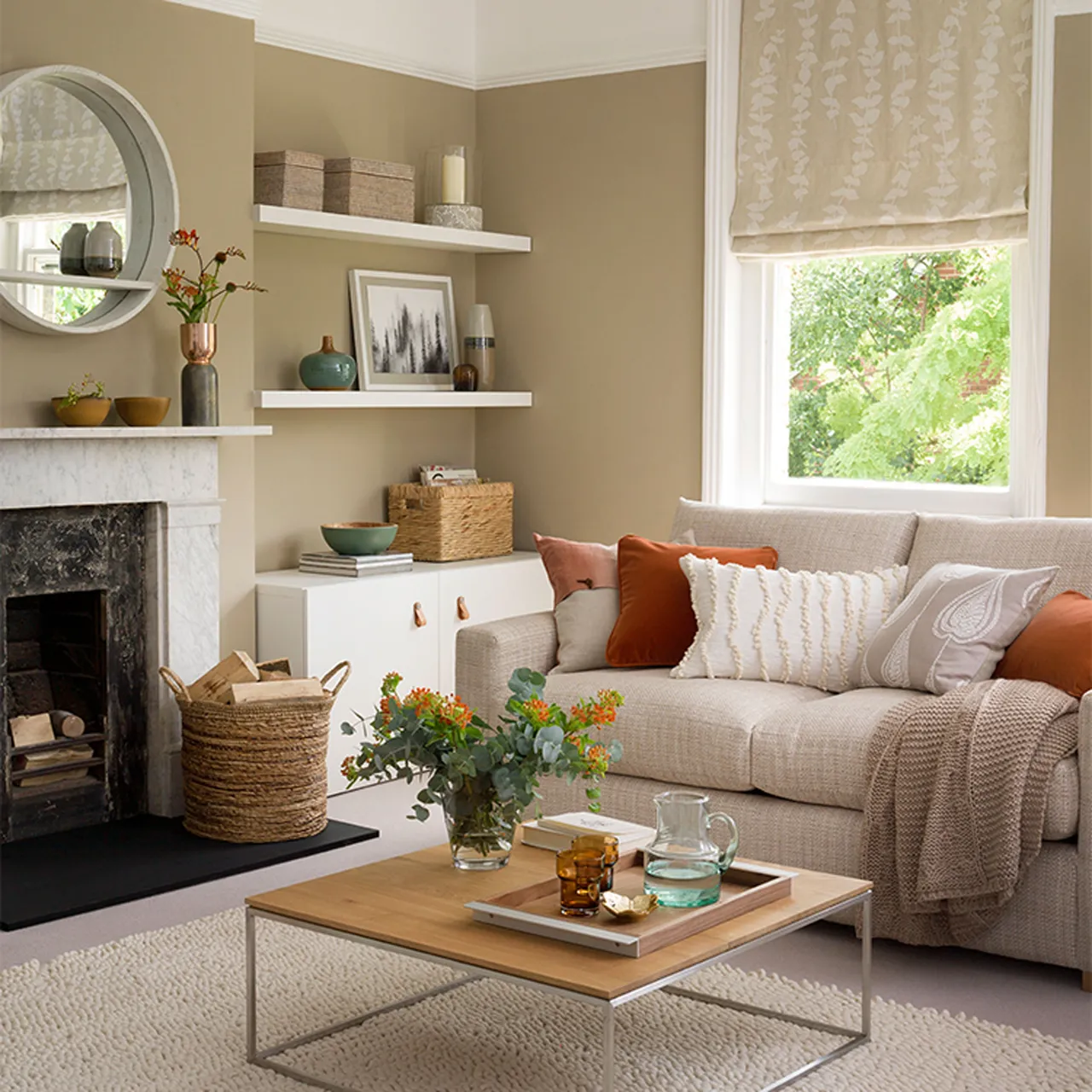 A cream-beige sofa has rust cushions beside a white fireplace and warm neutral walls