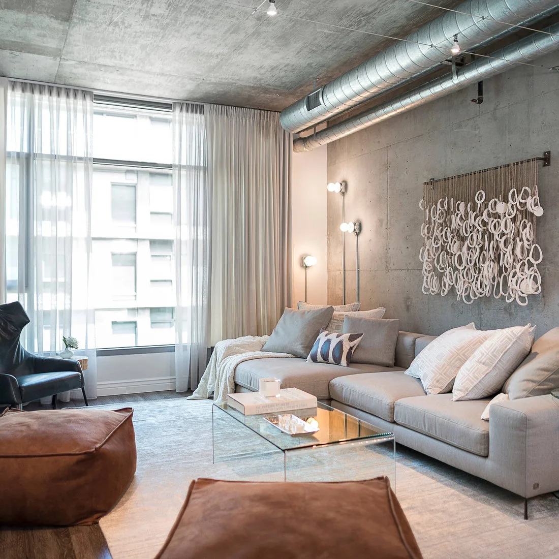 A pale sofa, brown ottomans and a looped wall hanging beneath concrete and silver ductwork