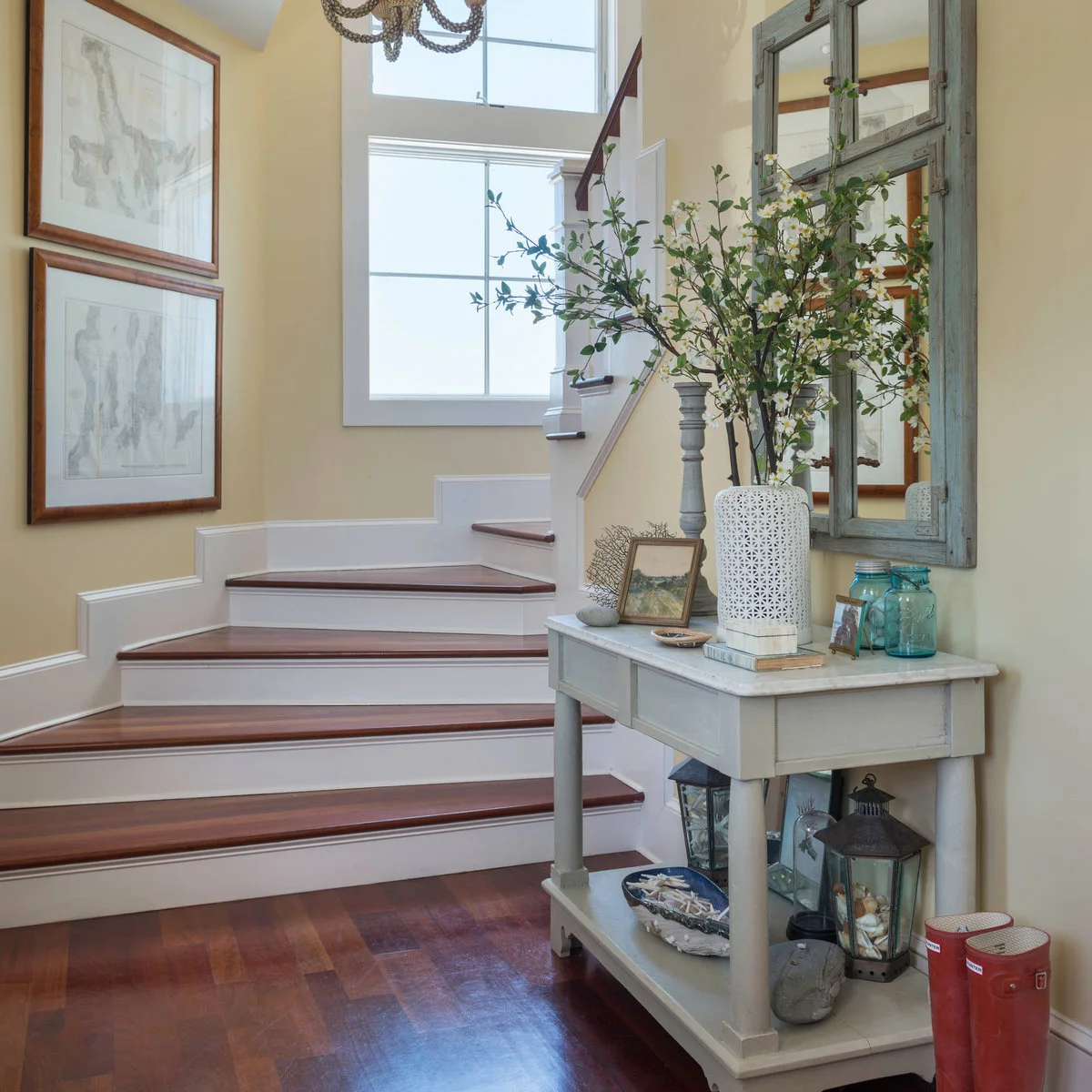 A pale two-level console beside stairs with a tall flower vase, small pictures, jars and lanterns on the lower shelf.