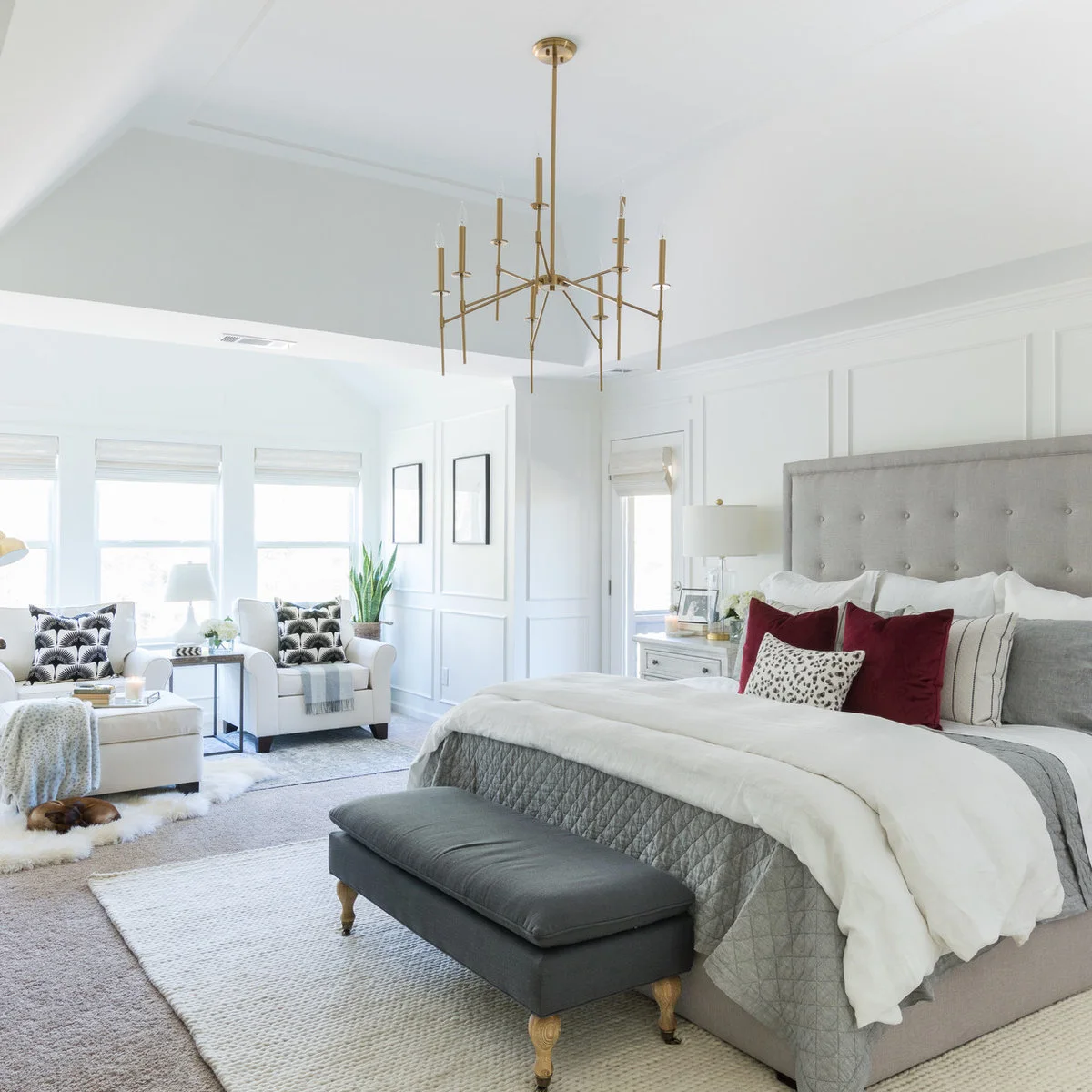 A cream rug lies over pale carpet beneath a gray bed and dark bench, with a separate sitting area by the windows.