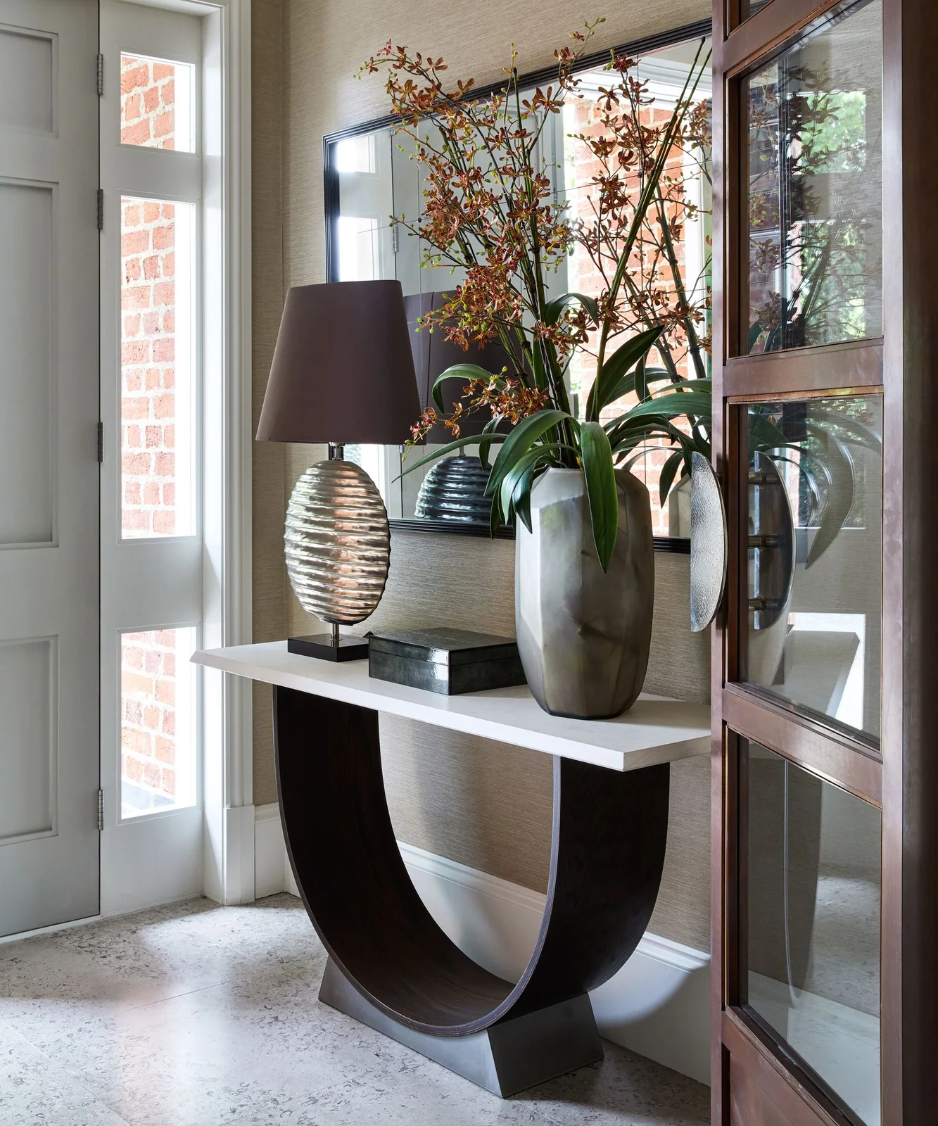 A pale console top rests on a dark curved open base beside a glazed door below a broad mirror with a shaded lamp and tall plant arrangement
