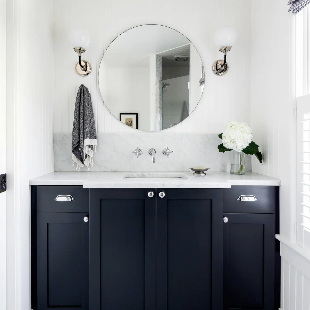 Narrow bathroom vanity with dark cabinet doors, a large round mirror and two wall lights