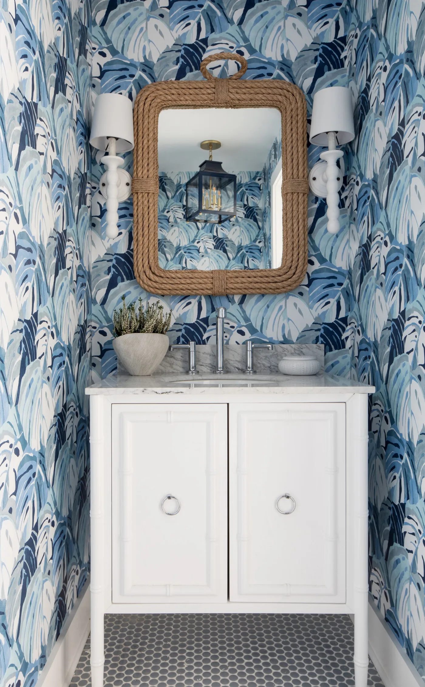Compact powder room with blue leaf-pattern wallpaper, white vanity and rectangular woven-framed mirror
