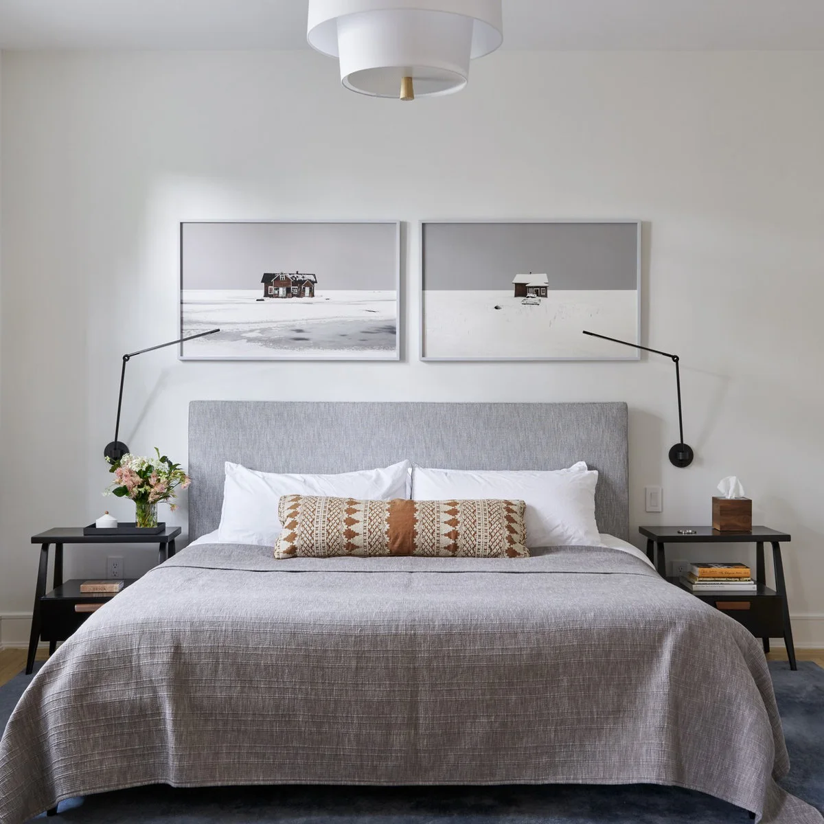 Two snowy landscape photographs above a gray headboard between slim black adjustable reading lights.