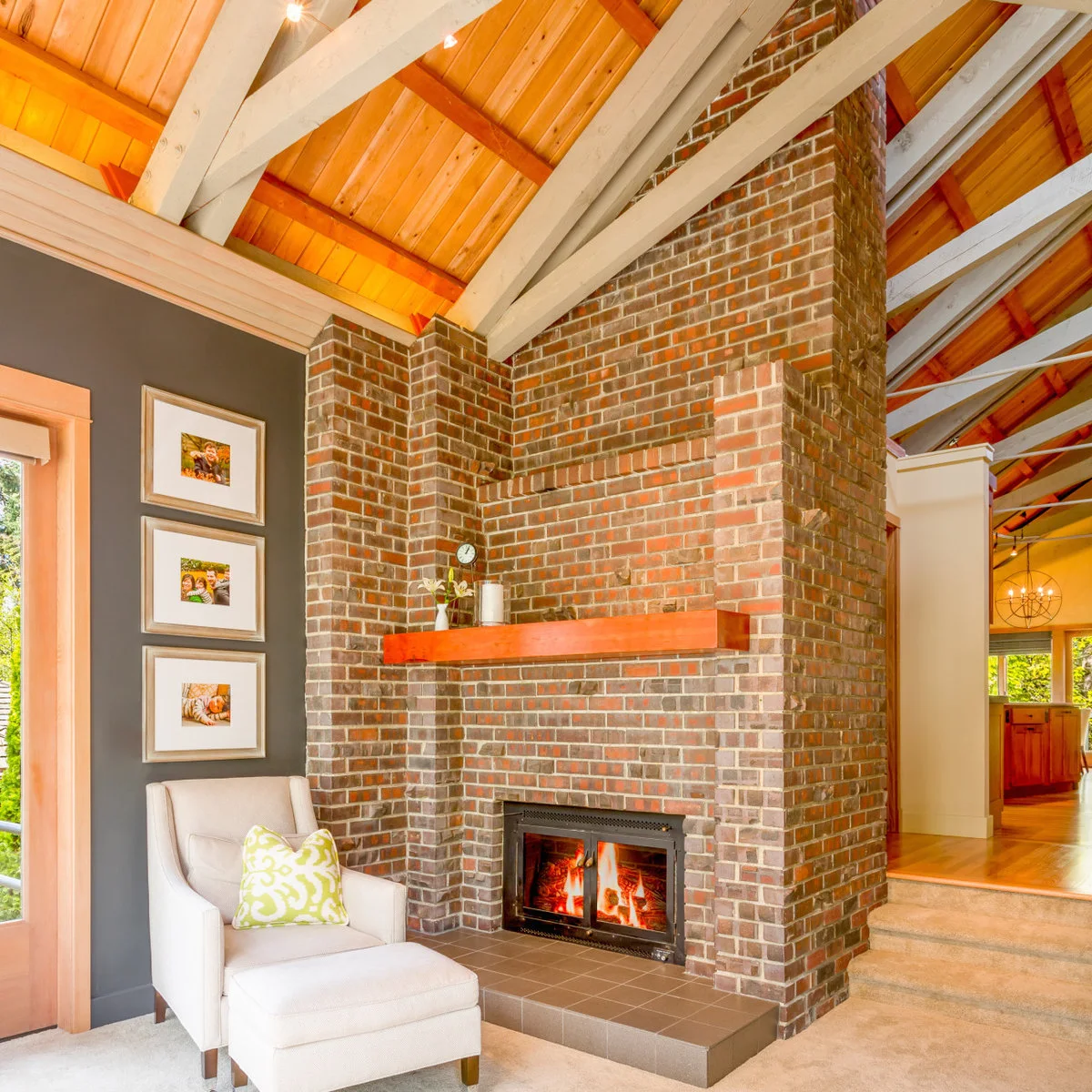 A tall mixed red and brown brick chimney rises beside white trusses and a wood ceiling, with a thick wood mantel and pale chair.
