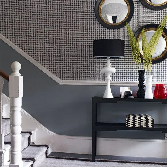 A hallway has small black-and-white patterned wallpaper above gray lower walls beside a dark console, black-shaded lamp and round mirrors