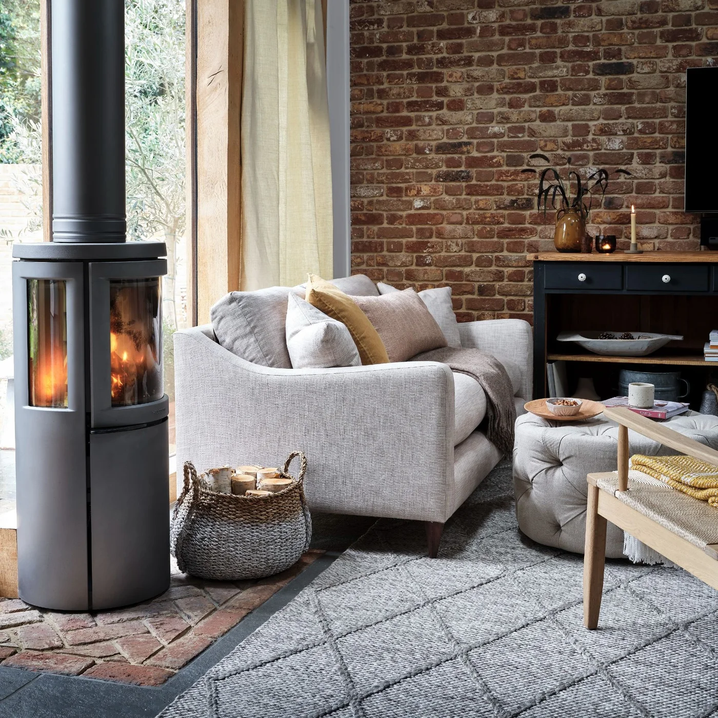 A pale gray sofa and tufted ottoman sit beside an exposed brick wall and a freestanding stove near broad glazing