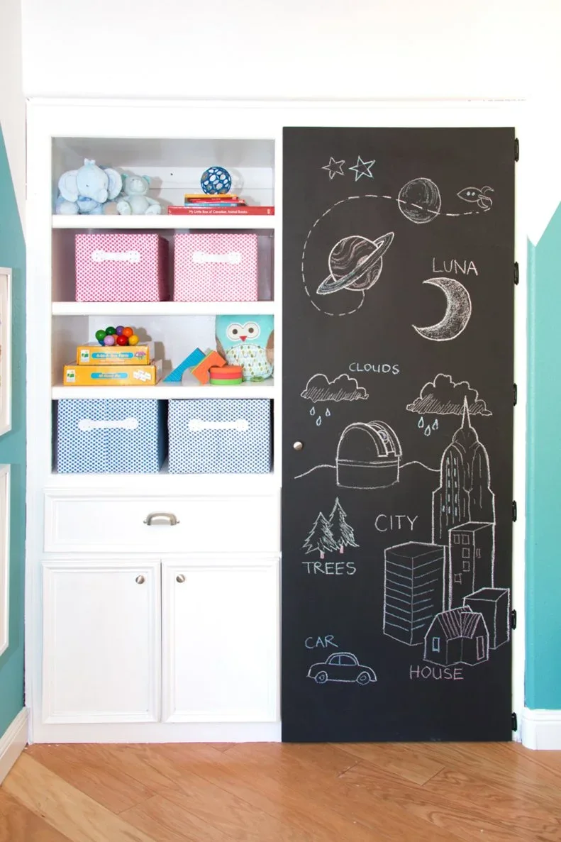 A white storage unit has shelves with patterned bins, closed lower doors and a tall black chalkboard door covered with drawings.