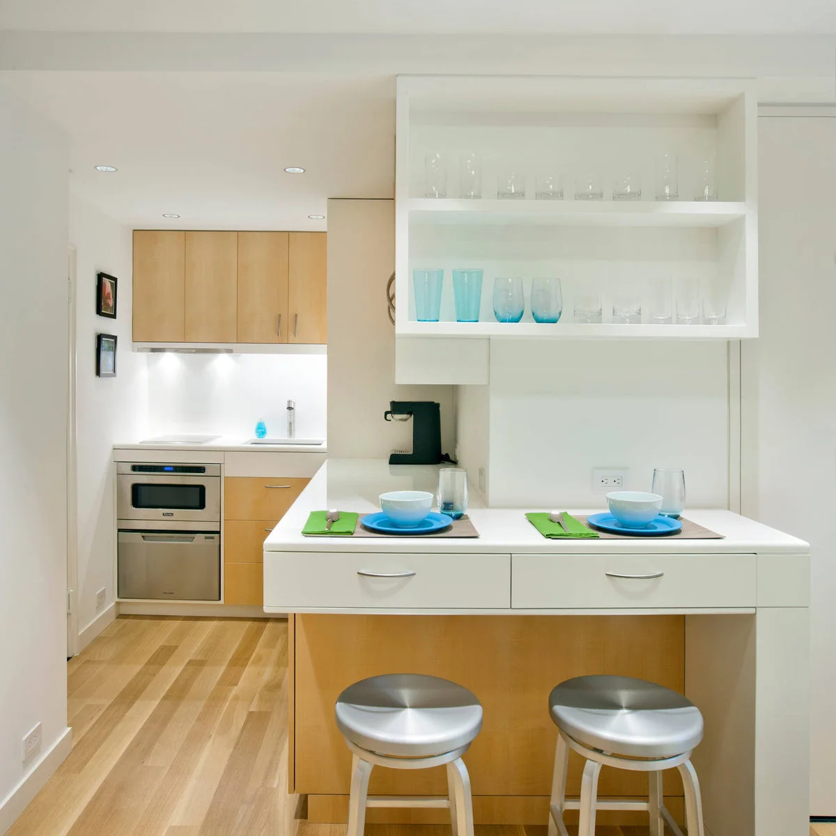 Compact kitchenette behind a white eating ledge with shallow drawers and two stools
