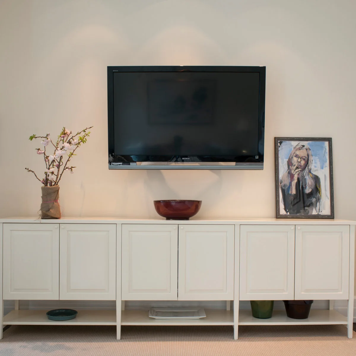 A flower arrangement, low brown bowl and framed portrait are spaced across a long white cabinet beneath a television.