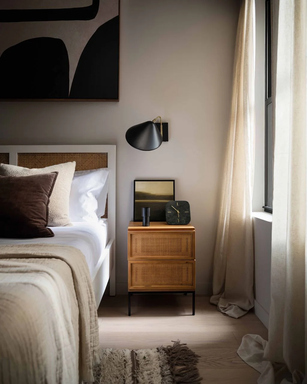 A pale bed with a woven-inset headboard stands beside a wood bedside chest and dark wall lamp beneath abstract artwork near light curtains