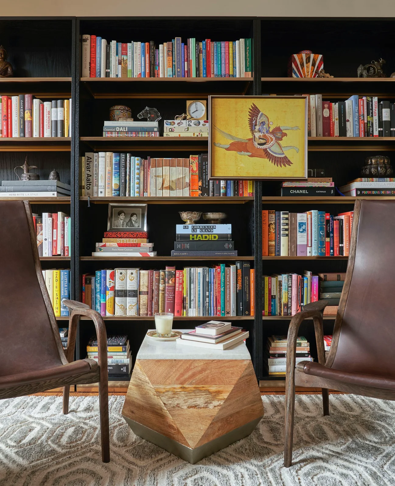 Dark bookshelves with wooden shelf edges hold books and framed artwork behind two brown chairs and a geometric central table