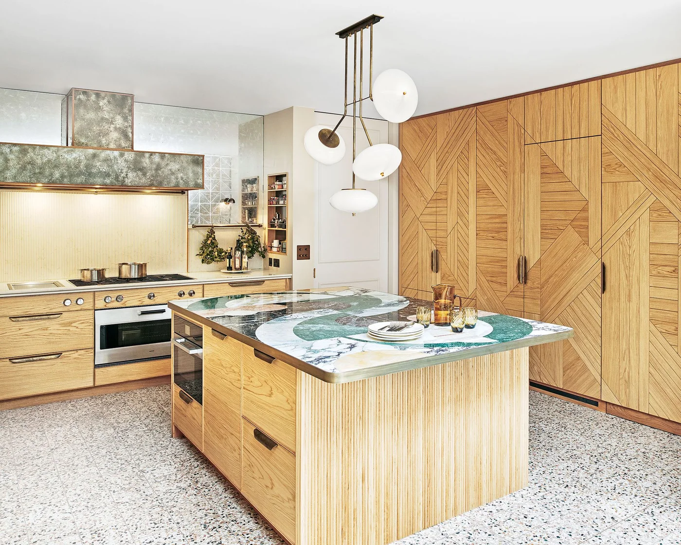 Geometric oak cabinet doors face a curved ribbed island with a green patterned counter, beside a metallic hood and pale backsplash.