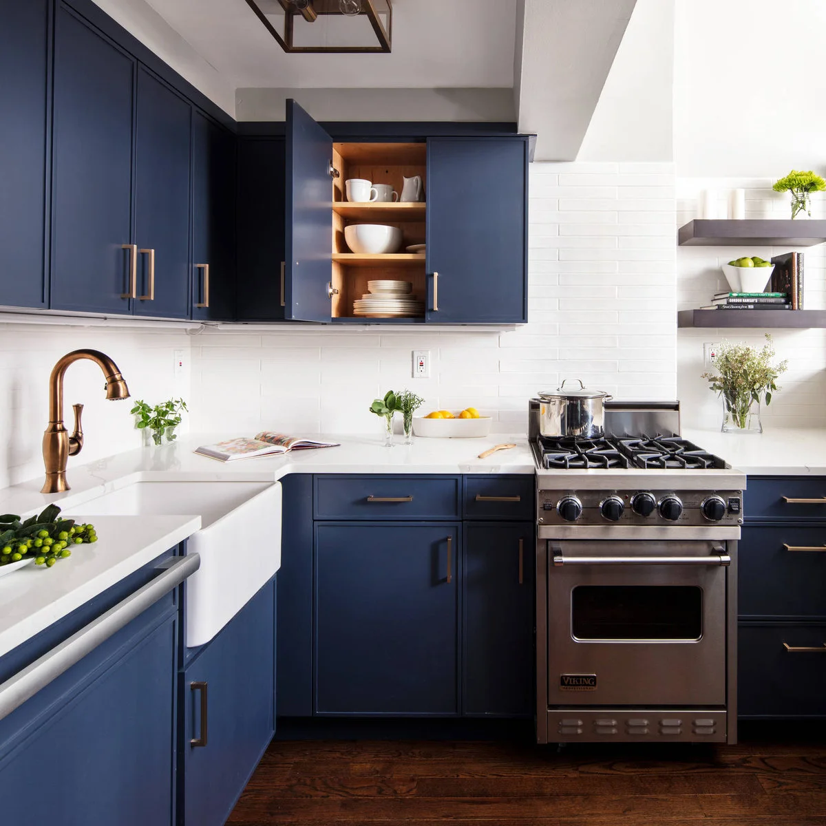 Navy upper and lower cabinets surround a white sink and counter, with white tile, warm metal fittings and dark wood flooring.