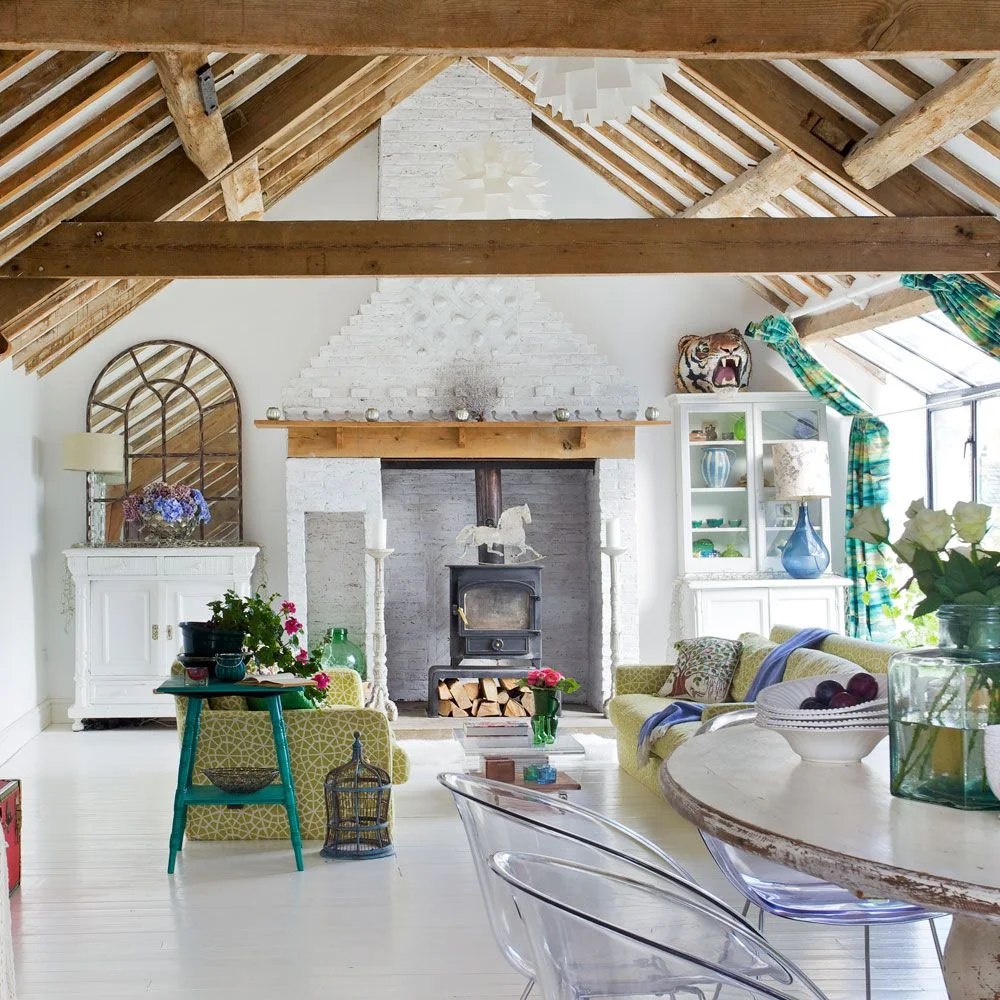 Green-patterned seating sits below a high timber roof beside a white fireplace and transparent dining chairs