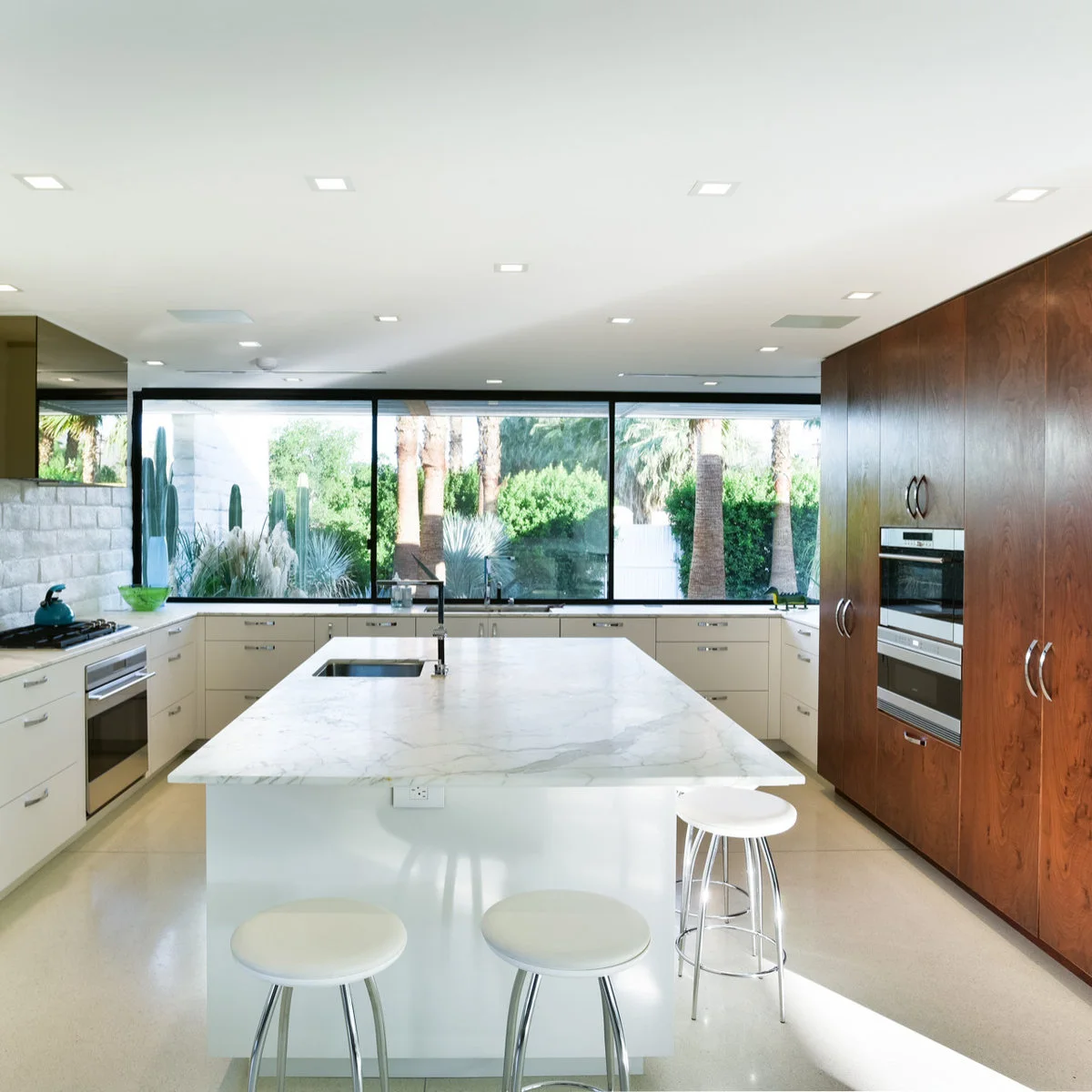A pale kitchen with a large garden-facing window, wood appliance wall and a long white island with simple stools.