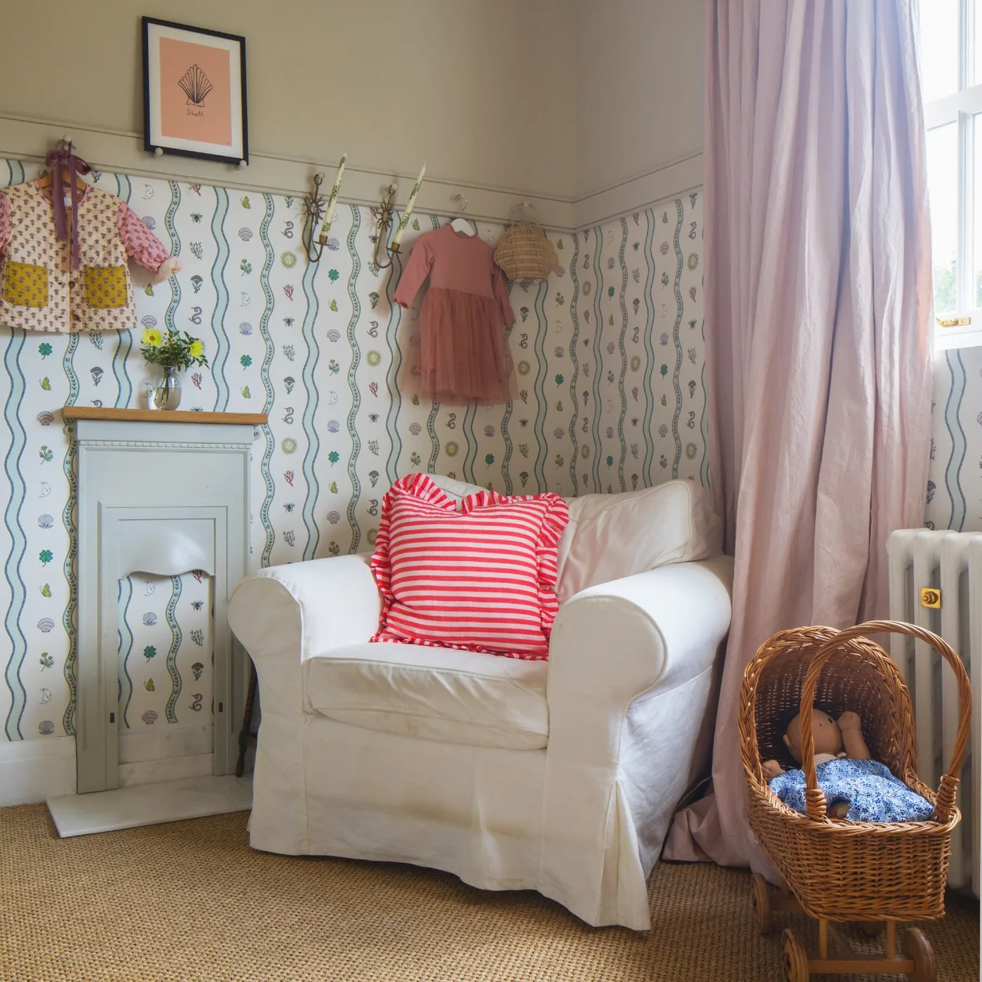 Pale slipcovered armchair with red-and-white striped cushion beside small fireplace, patterned lower wall and pink curtains.