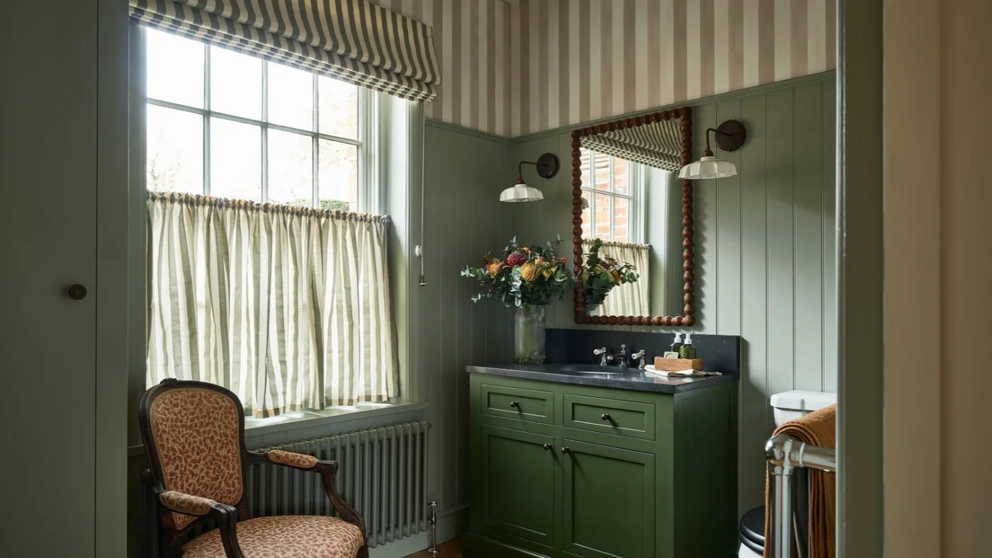 A green-paneled powder room has striped upper walls, a striped blind and cafe curtain, a wood-framed mirror and a small patterned upholstered chair.