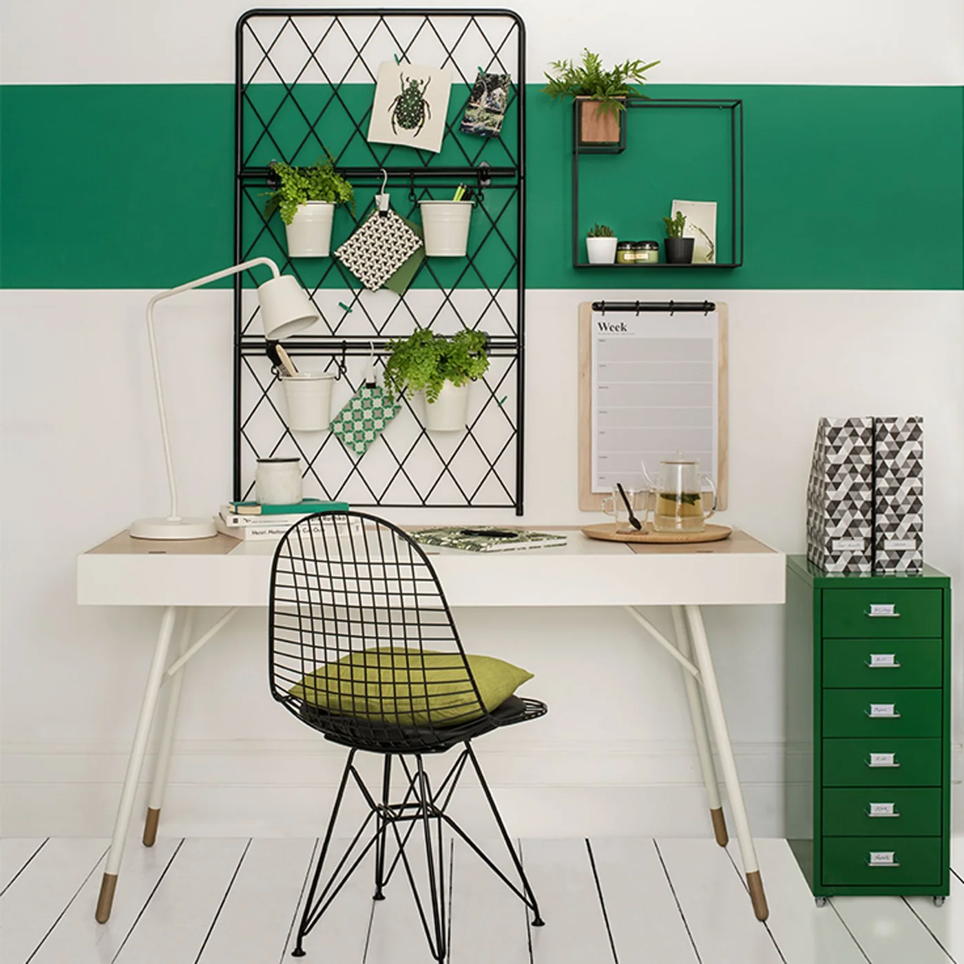 A green horizontal wall band sits behind a pale desk, dark mesh display panel and small green drawer cabinet
