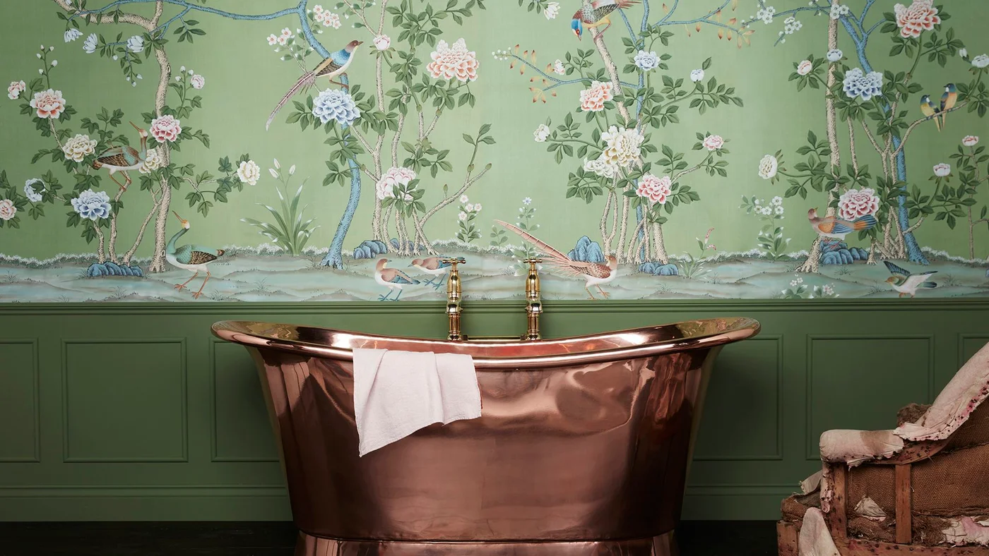A copper-colored reflective bath stands before green paneling and botanical wallpaper beside a patterned upholstered chair