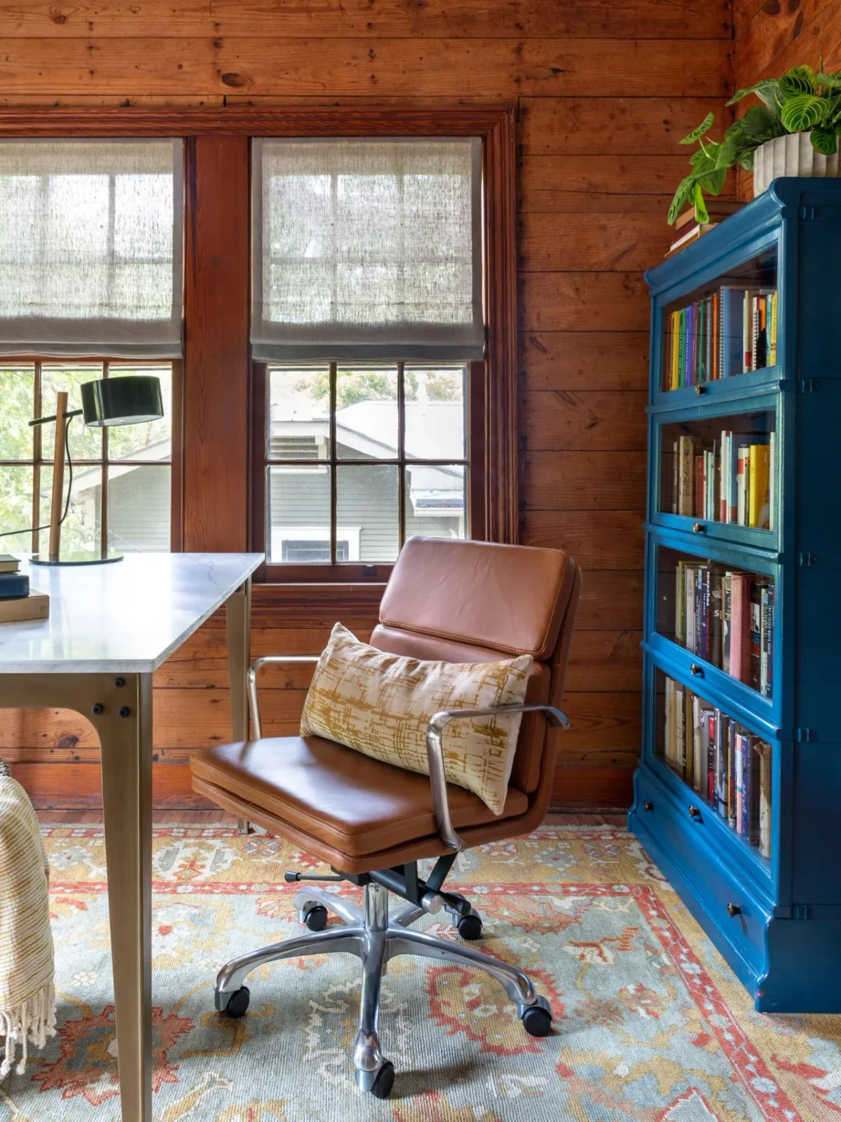 A wood-paneled office has a pale-topped desk, a brown rolling chair, a blue glazed bookcase, window blinds and a patterned rug.