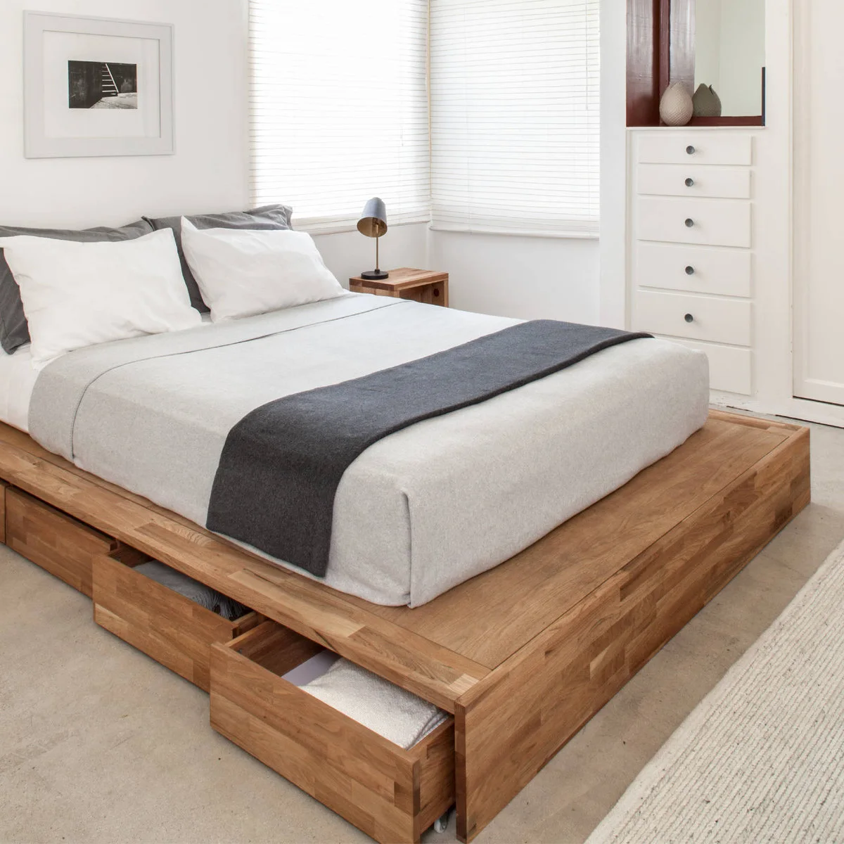 Wood platform bed with an open side drawer and a tall white built-in chest beside corner windows