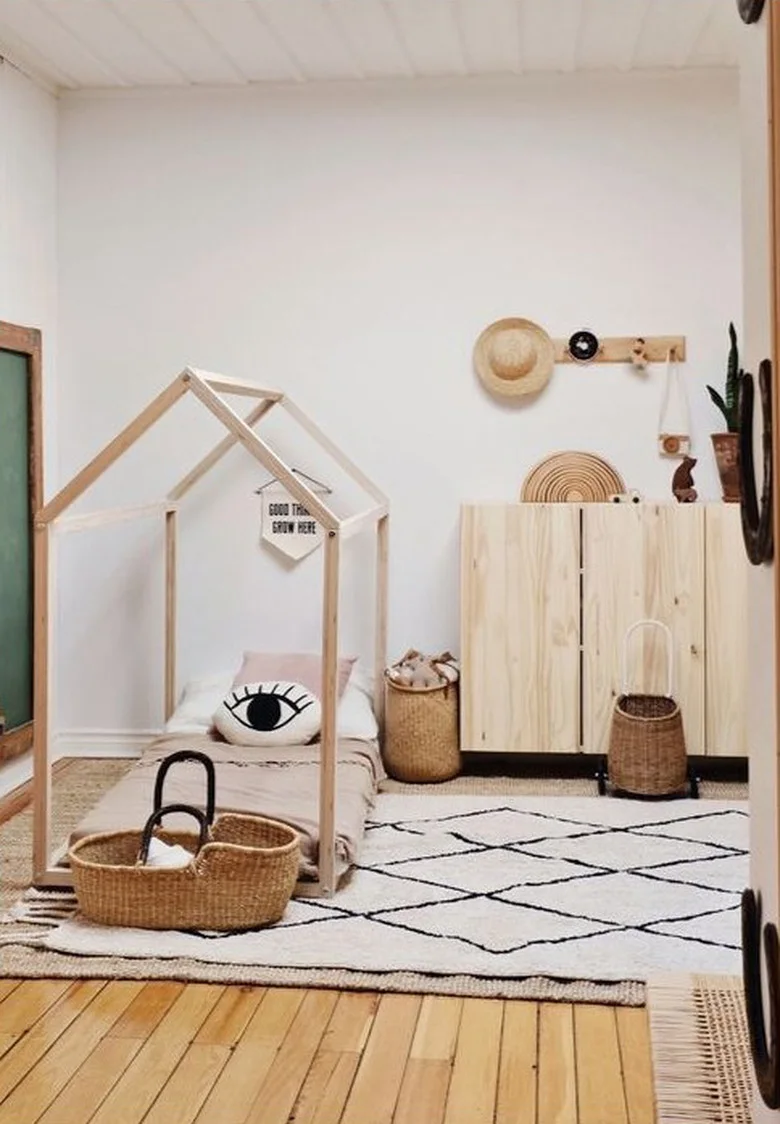 A house-shaped pale wood bed frame stands beside a plain wood cabinet, woven baskets and a light rug with black geometric lines